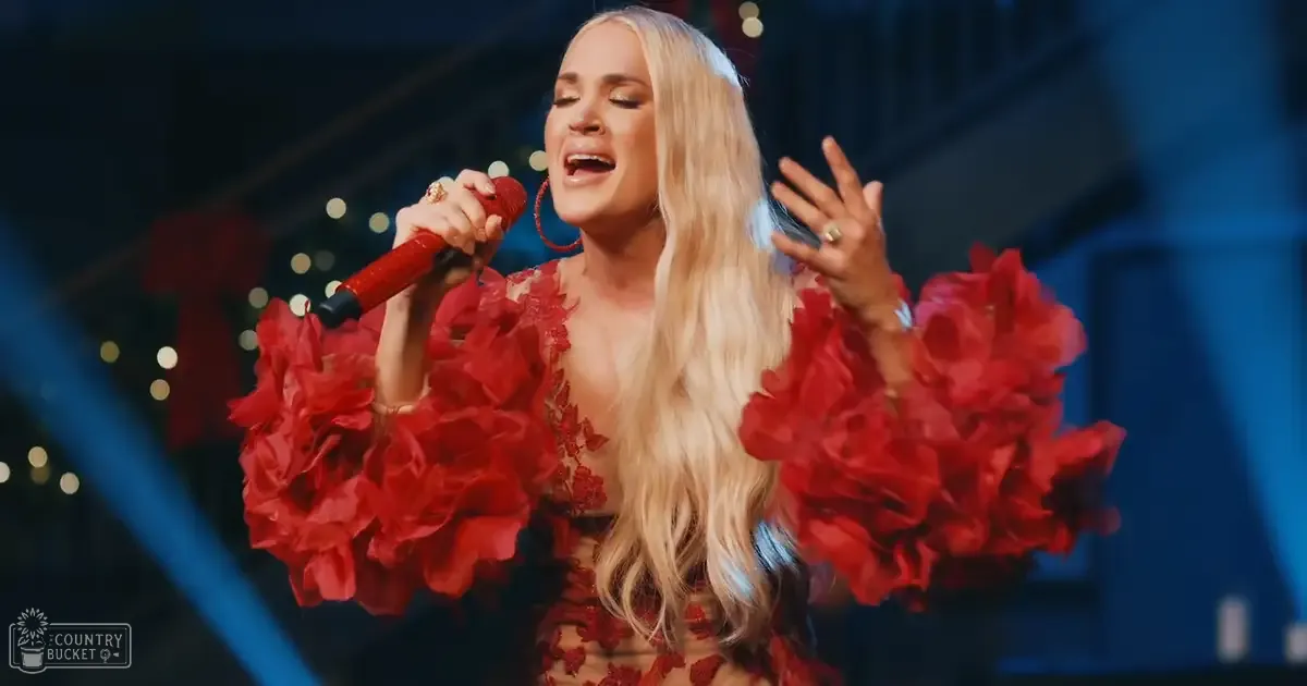 Adult female performer singing in a sleeveless red floral dress with form-fitting cut, deep V-neckline, large hoop earrings and rings, holding a red microphone on stage at an indoor concert venue with bokeh lights suggesting Christmas season.