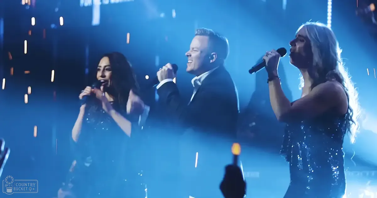 An indoor performance scene with microphones, blue stage lights, confetti, young adult female in a shiny dress on left, adult male in a dark suit jacket center, and another young adult female in a similar dress on right, modern event setting.