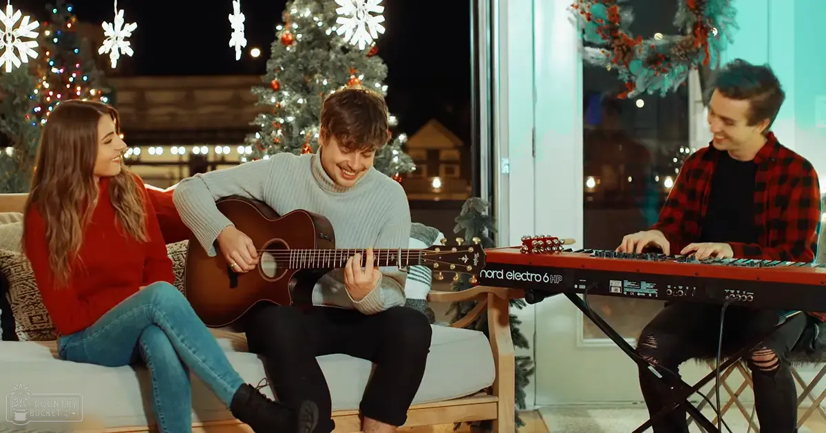 Young adult female in red sweater, young adult male in light gray turtleneck, and another young adult male in red plaid flannel shirt perform a musical Christmas concert indoors with white couch, Nord Electro 6 HP keyboard, and festive decor.