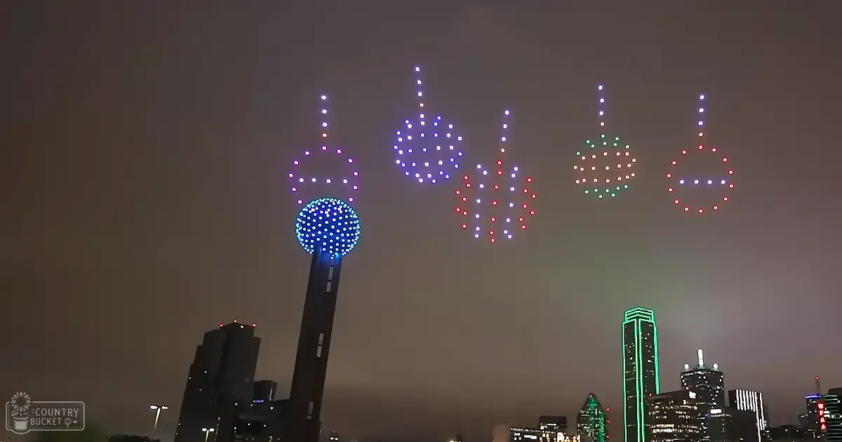 An urban nighttime scene with drones displaying colorful lights against silhouetted tall buildings in an illuminated city skyline.