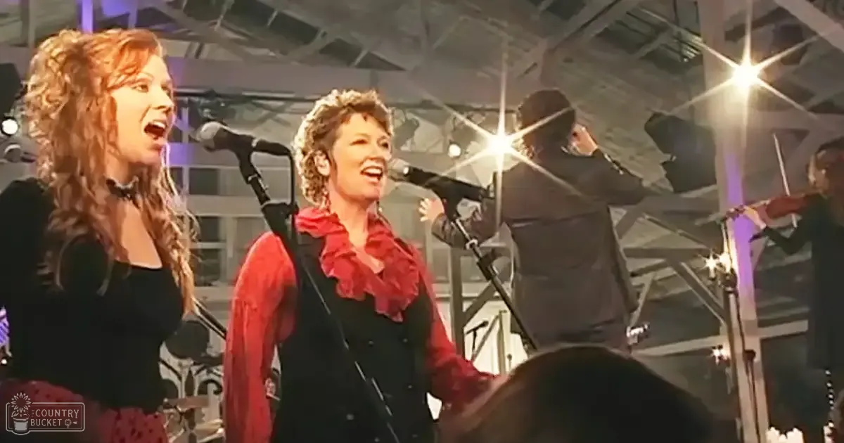 An adult female singer in a black top with red patterned skirt, accompanied by another adult female in a red scarf and dark vest, and an adult male in a dark suit jacket. The setting appears to be an indoor venue with industrial-style ceilings and lighting equipment.