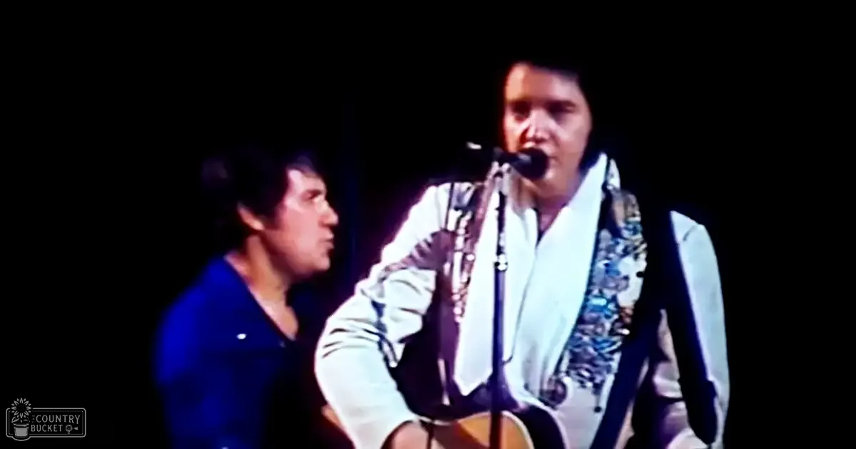 An adult male singer in a dark blue jacket, holding a microphone on stage during what appears to be a concert or performance with another adult male performer wearing a white patterned shirt and holding an acoustic guitar.