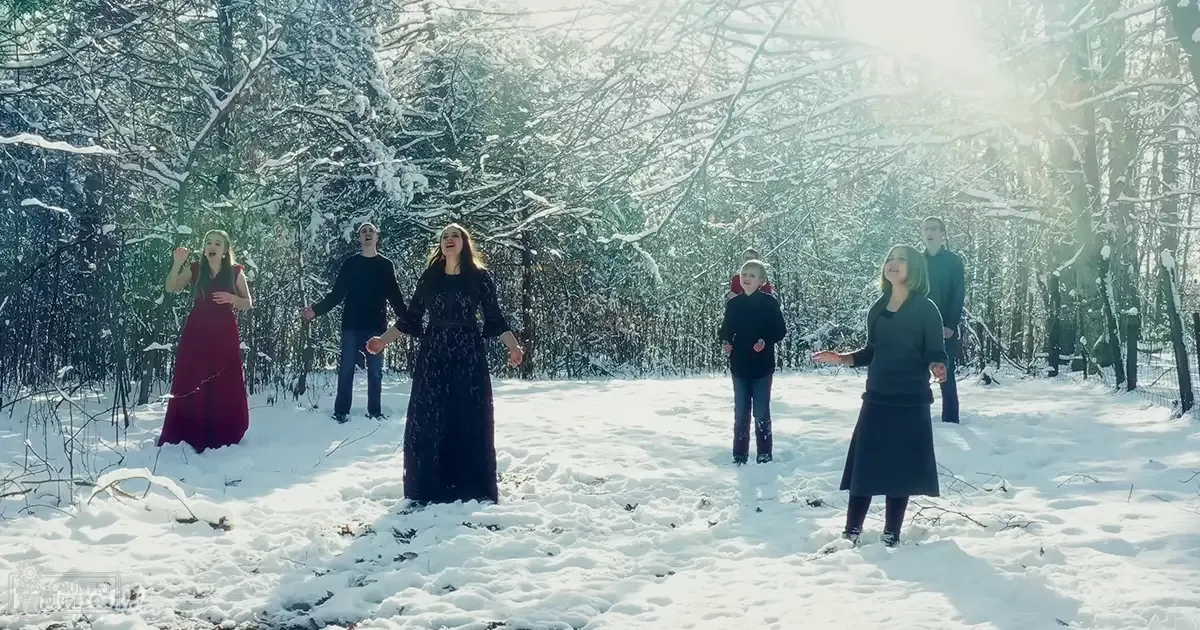 A group of people singing in a snowy forest with bare trees, sunlight filtering through branches at late afternoon or early evening.
