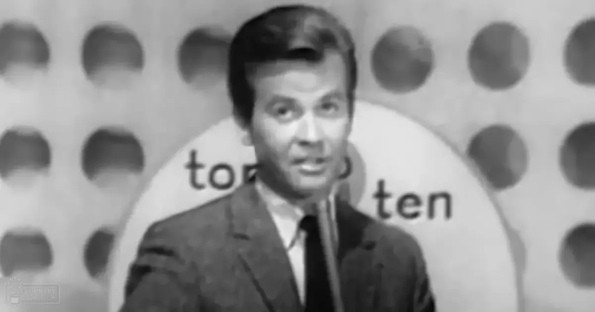 An adult male wearing a dark suit jacket and white shirt stands before a 'Top Ten' sign in an indoor event setting from the mid-20th century.