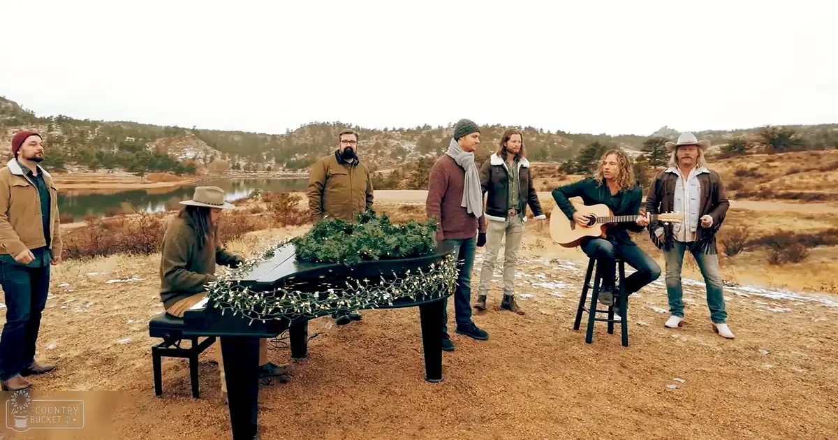 A musical performance with a piano decorated with lights and greenery set against an outdoor backdrop with dry grass, shrubs, hills, and a body of water in the distance.