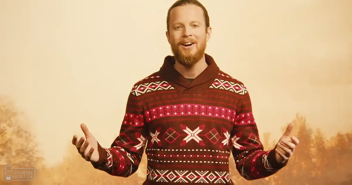 A young adult male wearing a red Fair Isle hoodie with white and pink geometric patterns, indoors in an autumn setting with warm lighting.