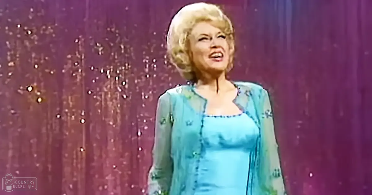 A middle-aged woman in a turquoise dress with small decorative patterns performs on stage, set against a purple curtain with sparkling embellishments.