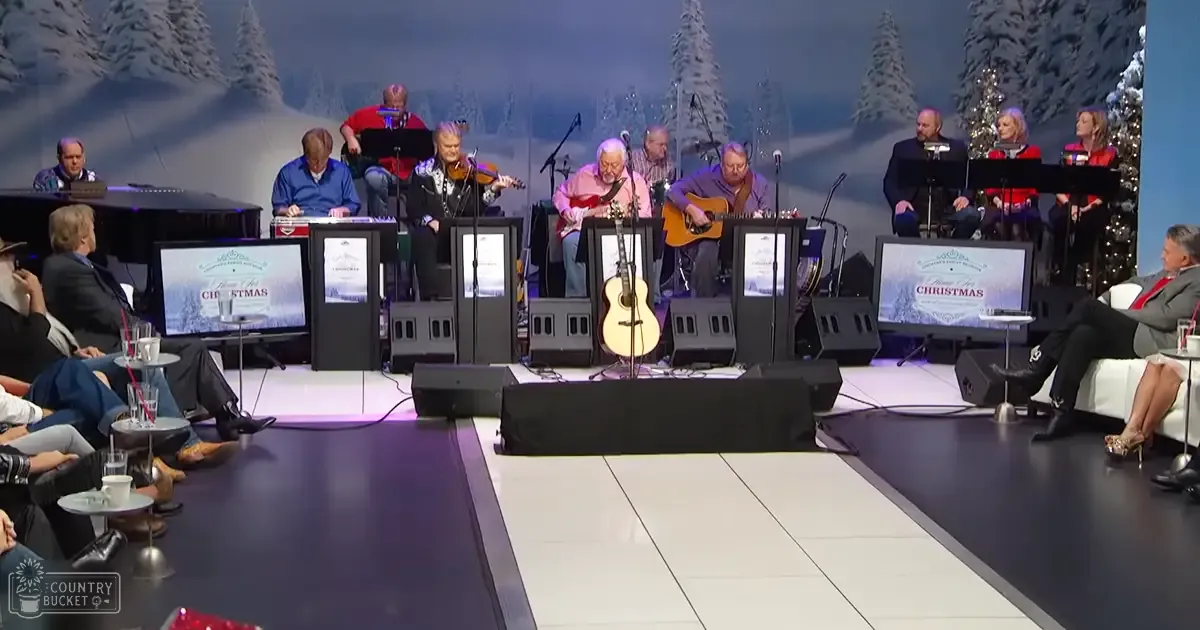 A musical performance during Christmas time with a snowy backdrop, multiple performers on stage, and bright indoor lighting.