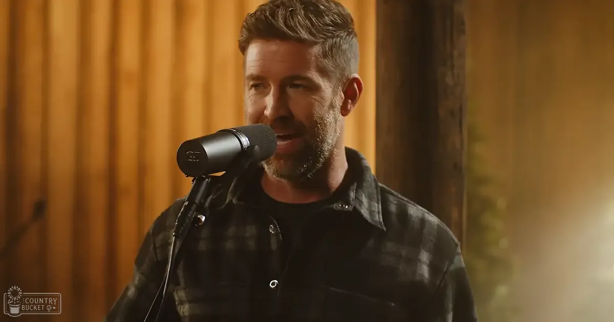 A middle-aged adult male in a casual plaid shirt, singing or speaking into a microphone in an indoor studio setting with wooden paneling and warm lighting.