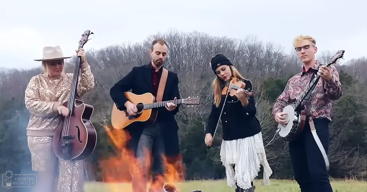 A small group performing music outdoors near a campfire, dressed in vibrant outfits with a rural autumnal backdrop.