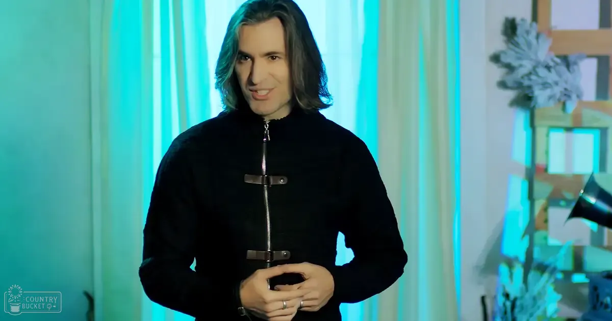 A young adult male stands indoors in a studio setting wearing a black zip-up jacket with two horizontal buckles across the chest, surrounded by holiday-themed decor and vertical light strips.