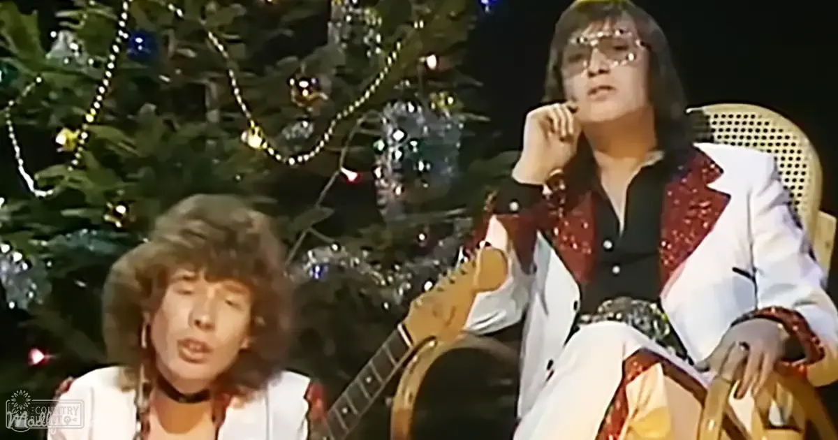 Two young adult males perform on stage; one wears a white jacket with red accents and black choker, another in a similar outfit with sequins, against a decorated Christmas tree backdrop.