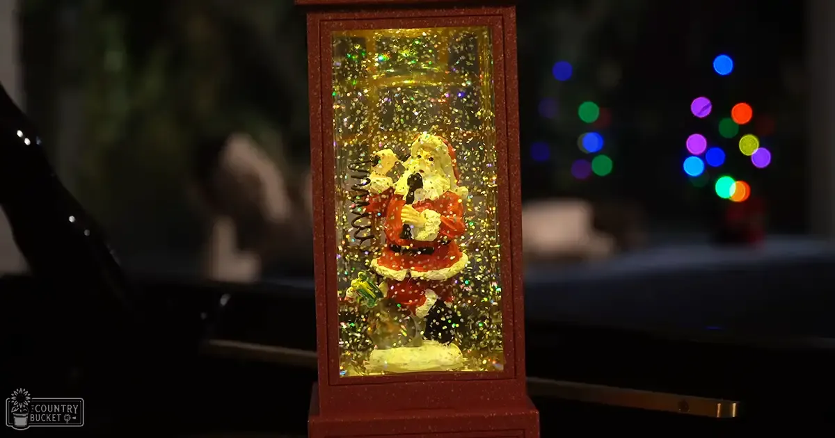 A decorative Santa Claus figurine inside a red glittery lantern, set against blurred Christmas lights in an indoor holiday scene.