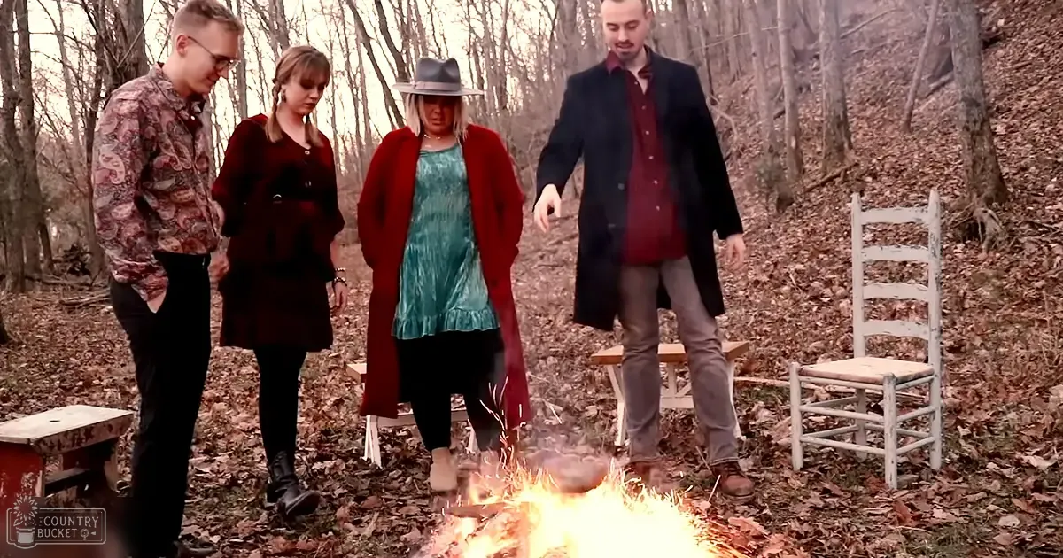 Four adults outdoors in a wooded area during autumn; young adult male with patterned shirt and glasses, adult female in dark red coat, middle-aged female in teal dress with wide-brimmed hat, adult male in blue coat near wooden chair and campfire.