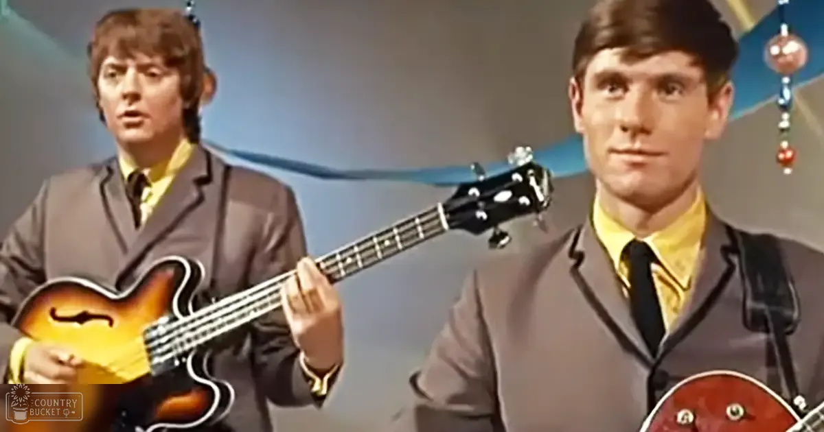 A man in a suit plays a bass guitar, another man in a suit looks on, both in a studio setting.