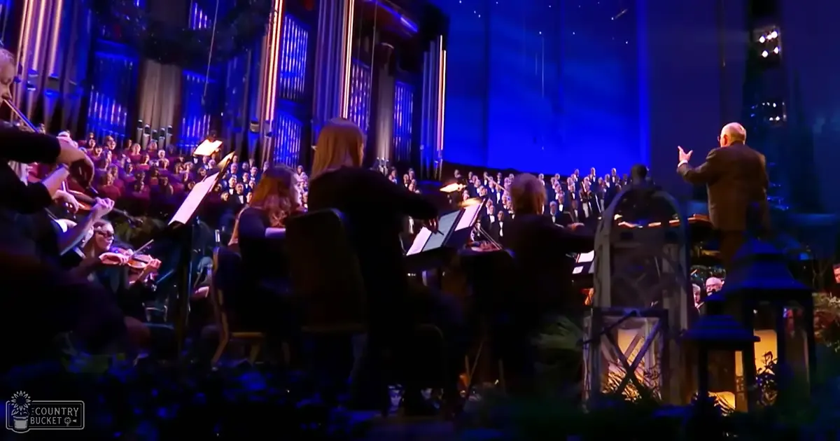 A live orchestral performance in an indoor concert hall with musicians wearing dark formal attire and a conductor in a dark suit jacket, ornate architectural details, and stage lighting predominantly with shades of blue.