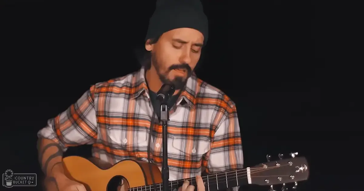 An adult male musician wearing a black beanie, plaid orange, white and gray striped shirt with button-down collar and short sleeves, performing in a dimly lit indoor studio setting focused on the performer and their acoustic guitar.