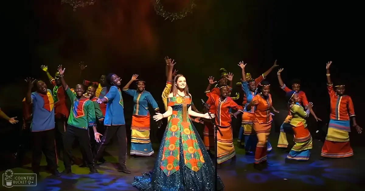 A young adult female performer stands center stage amidst a vibrant cultural event with colorful traditional African attire.