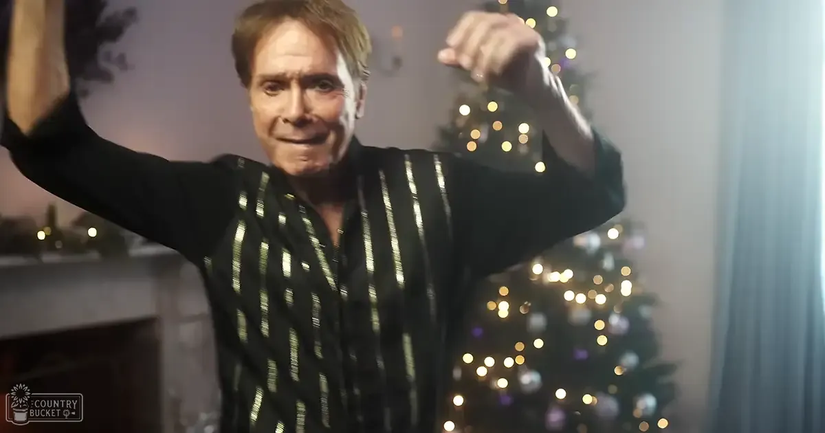 An older adult male dressed in a festive black shirt with gold stripes stands indoors during Christmas time near a decorated tree and fireplace mantel adorned with small ornaments.