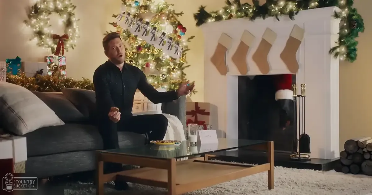 A festive living room decorated for Christmas with a young adult male wearing a patterned dark blue/black long-sleeved shirt, surrounded by holiday decorations including a wreath, stockings, and wrapped gifts.