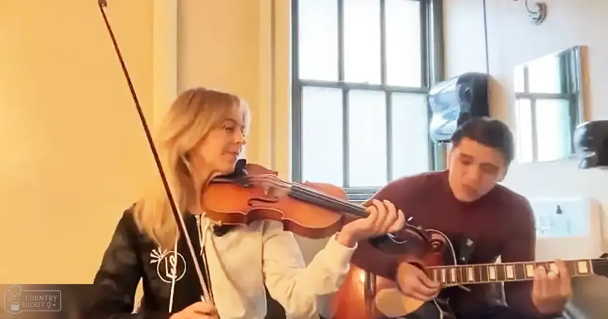 A musical performance with a female violinist in a white hoodie and black jacket, and a male guitarist in a dark long-sleeve shirt. The setting is an indoor room with large windows allowing natural light during what seems to be late morning or early afternoon.