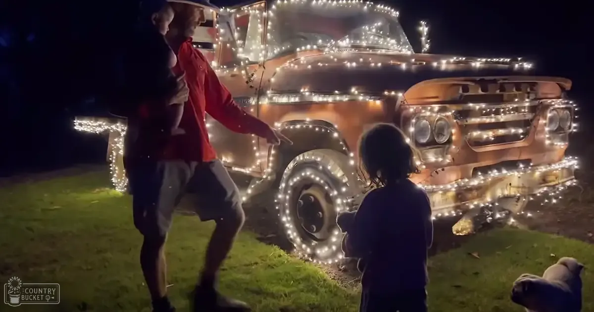 A nighttime outdoor family scene with a vintage truck decorated with string lights, two sheep partially visible, and grassy ground.