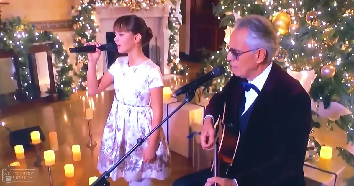 A young girl sings into a microphone with an older man playing guitar, surrounded by Christmas trees and festive decorations indoors.