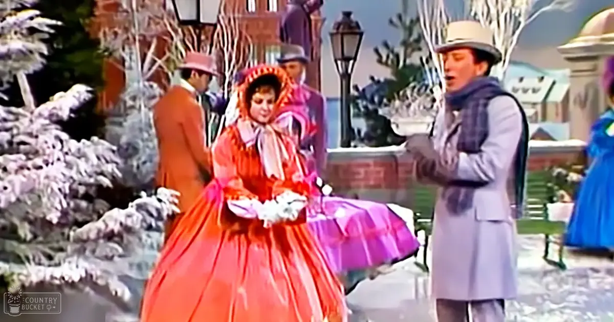 A woman in a red gown with puffed sleeves and a large bonnet, accompanied by an adult male in a light gray overcoat and hat, amidst snow-covered trees and vintage street lamps in what appears to be a historical outdoor setting.