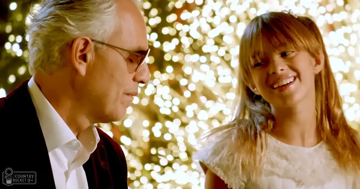A festive indoor scene with an older adult male and a child female in formal attire against out-of-focus holiday lights.