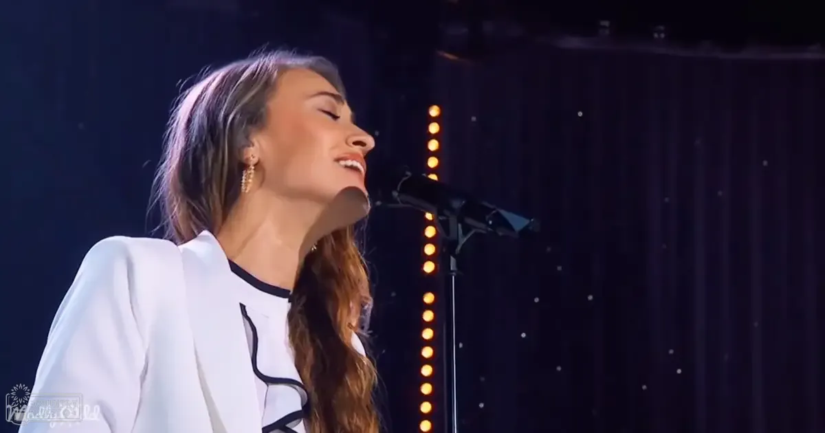 A young adult female singer wearing a white blazer with black trim and ruffles, performing into a microphone against an indoor stage setup with vertical light strips.