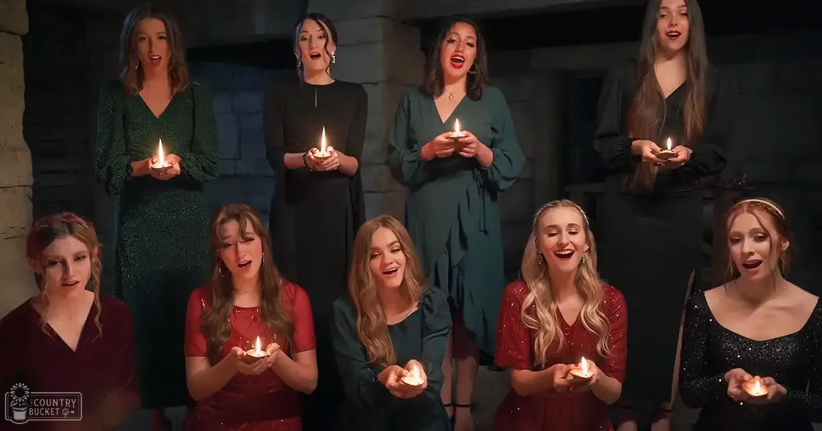 A group of young adult females singing in an indoor setting with stone walls, holding lit candles; dark reds, teals, and blacks dominate the scene.