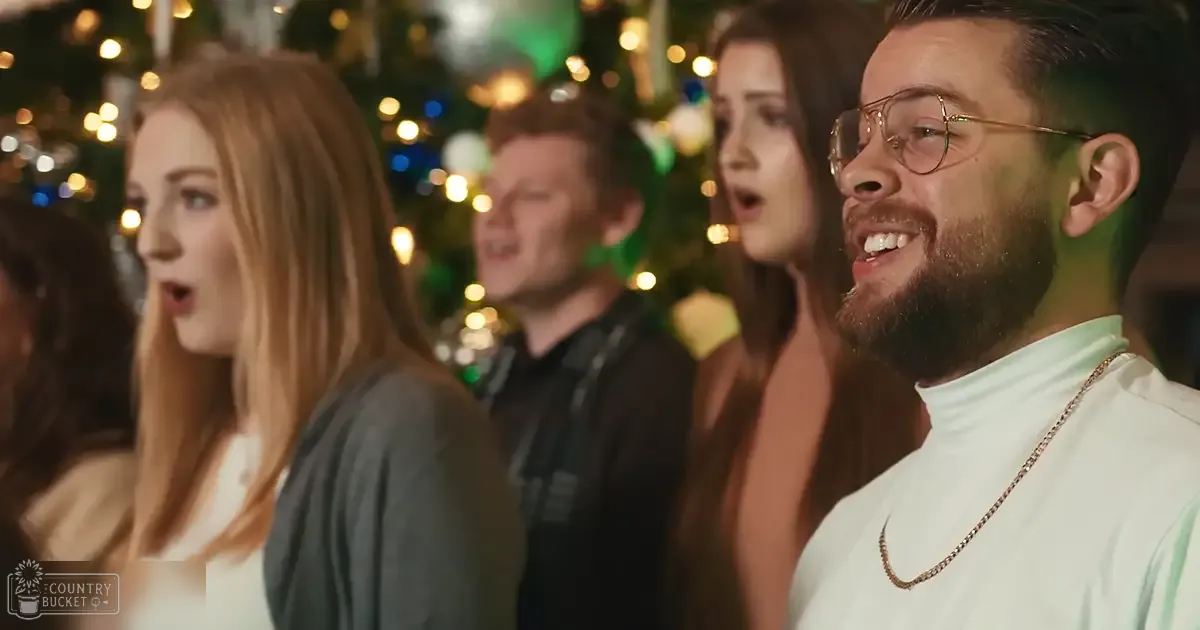 A group of adults singing indoors during a holiday gathering with Christmas lights and ornaments, suggesting evening or nighttime.