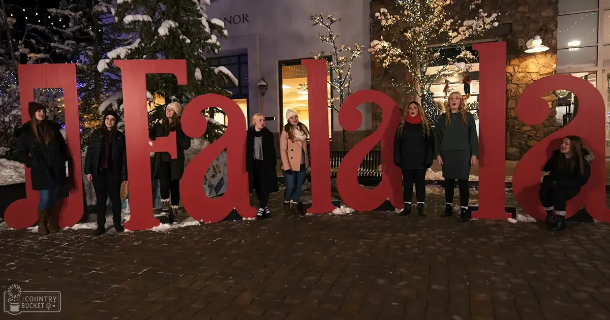Group photo in front of festive 'Fa la la' display with snow-covered trees, evening lights, and commercial setting.
