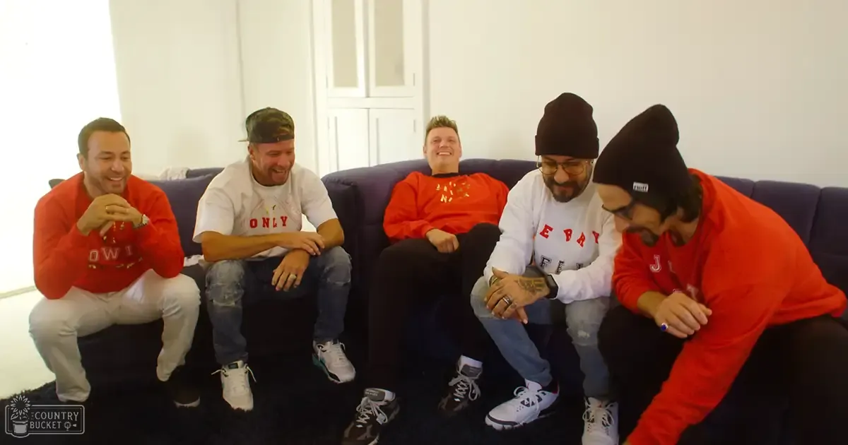 Five adult males sitting on a dark couch in an indoor living room with white walls, wearing various casual shirts and jeans.