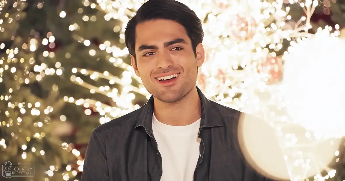 A young adult male in a relaxed fit, dark gray/black button-up shirt over a white t-shirt stands indoors amidst festive decorations, including a Christmas tree with white lights and red ornaments.