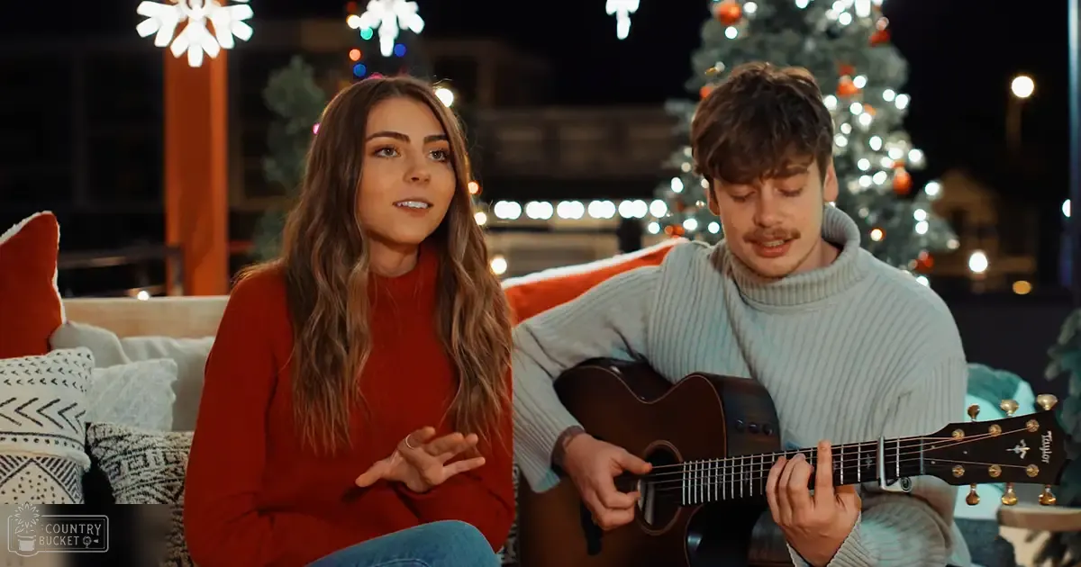 A young adult female with long brown hair wearing an orange turtleneck sweater and blue jeans stands next to a young adult male with short dark hair and a mustache, playing a guitar in a festive setting decorated for Christmas.