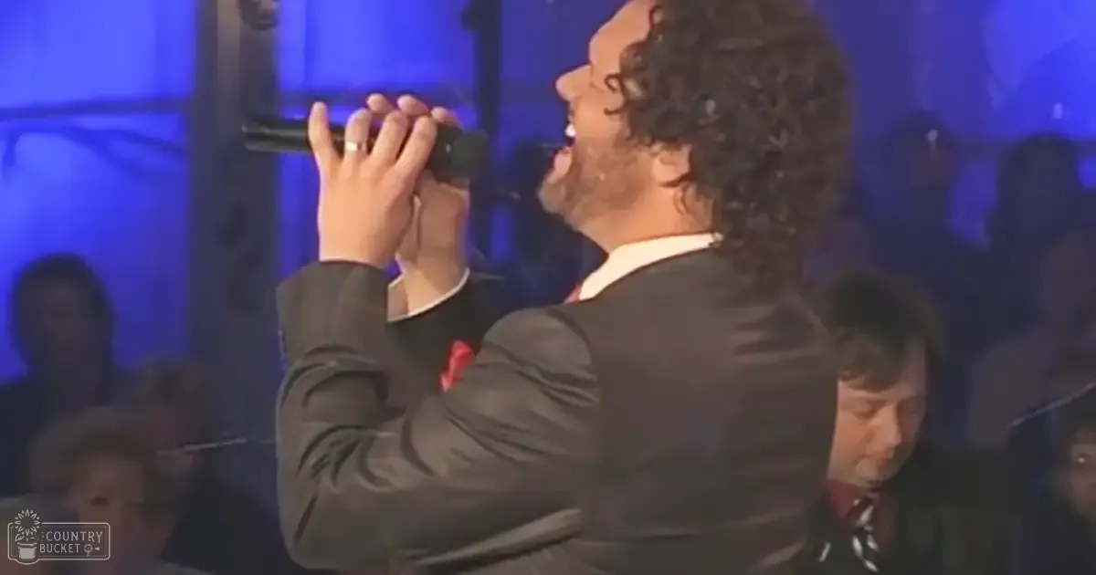 Adult male singer wearing a dark suit jacket, performing into a microphone at an indoor concert with blue stage lighting and seated audience in rows.