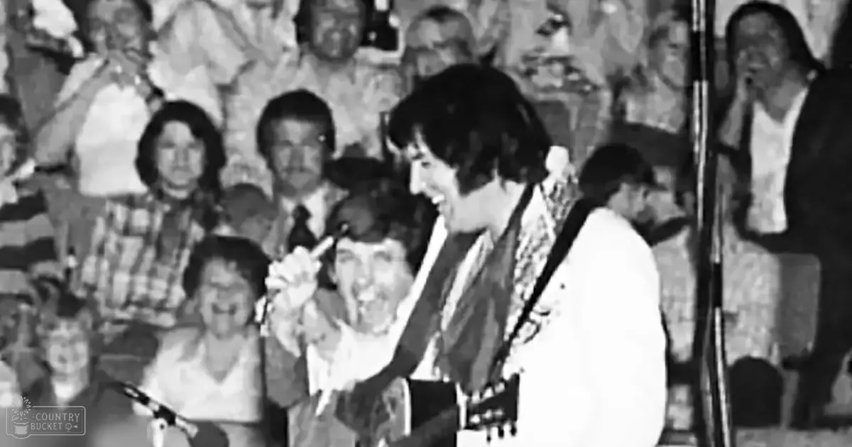 A young adult male performer in a patterned scarf and white shirt on stage at an outdoor concert venue, with audience members in casual attire seated behind him.
