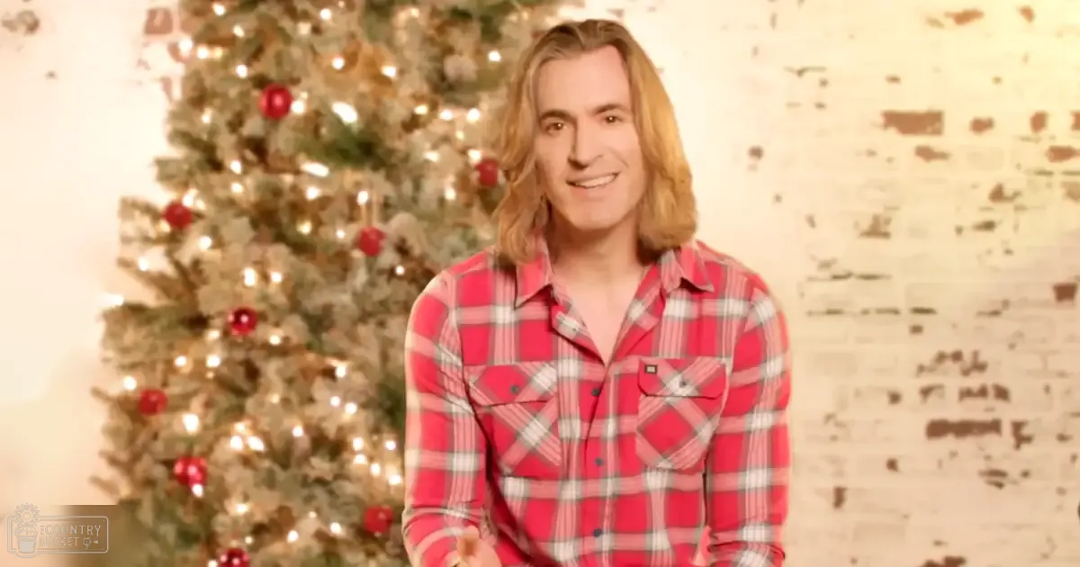 A young adult male in a red plaid shirt stands in front of a decorated Christmas tree against a textured brick wall, suggesting an indoor winter evening setting.