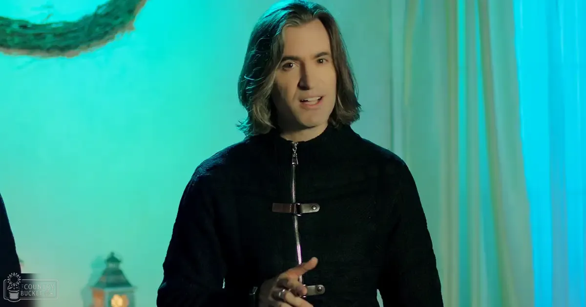 A middle-aged man wearing a black zip-up sweater with textured pattern and high collar stands indoors against a light teal background with vertical blinds. A faint outline of a clock tower suggests an urban setting, possibly during daylight hours.