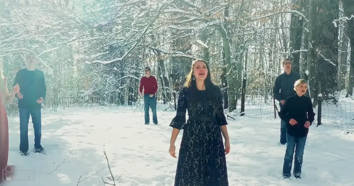 A young adult woman sings in a snowy forest scene with two teenagers on the left, an adult male on the right, and a child at far right. Snow-covered trees and a wire fence frame the setting.