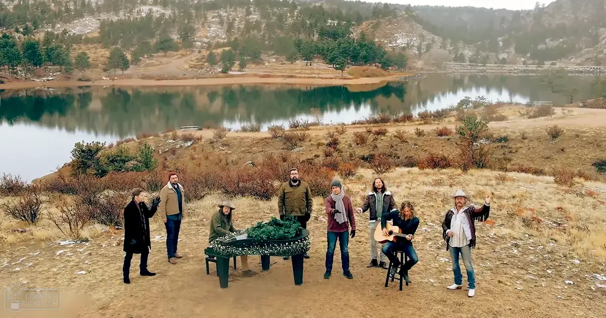 Group of musicians outdoors near a lake in autumnal setting with earthy browns and greens dominating the scene.