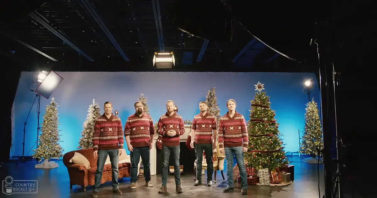 Group of five adult males in a studio setting for a holiday-themed photoshoot wearing red patterned V-neck sweaters with white crosses, dark jeans, and brown shoes. Background includes Christmas trees, lights, ornaments, couch, pillows, gift boxes, blue backdrop, and lighting equipment.