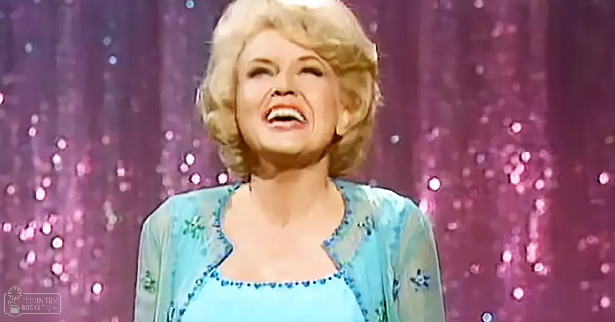 A middle-aged adult female wearing a loose-fitting, long-sleeved, light blue dress with embroidered floral patterns stands against a sparkly curtain in an indoor setting illuminated by artificial lighting.