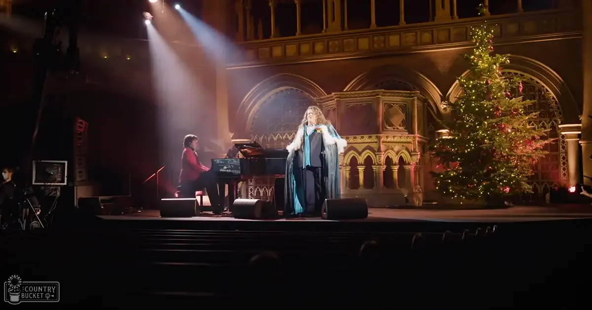 A performer in formal attire with a cape, standing on stage at what seems to be a concert venue during the holiday season.