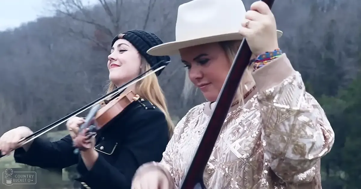 Two young adult female musicians playing stringed instruments outdoors in a natural setting with trees and hills in the background.