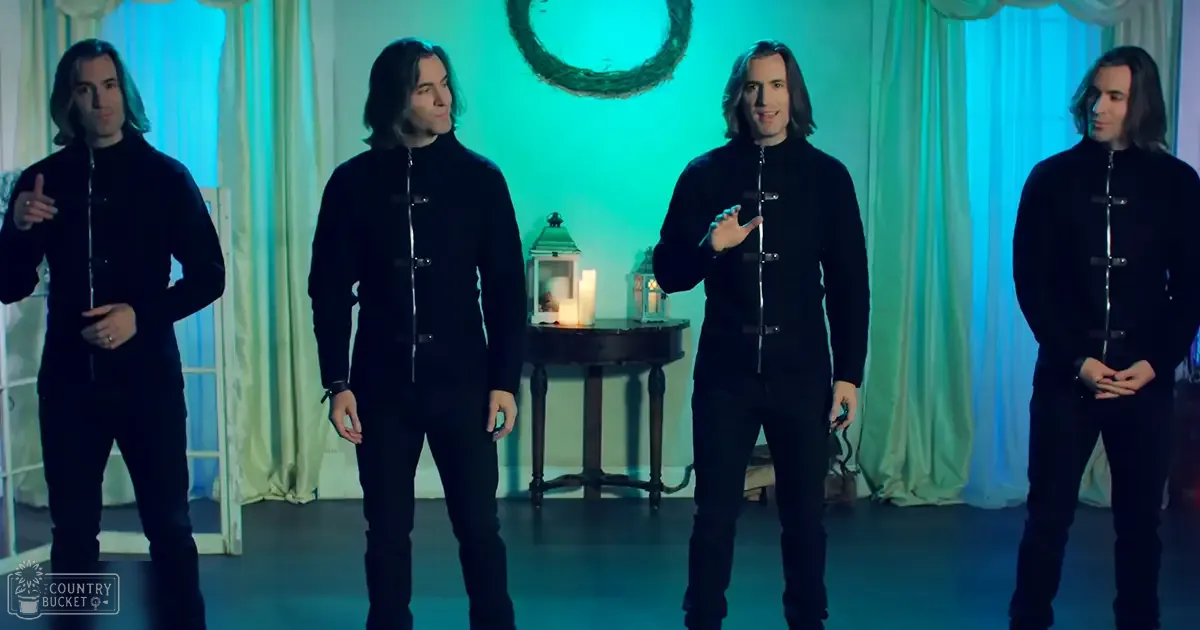 Group of adult male performers with long hair in an indoor studio setting. They wear black zip-up jackets and dark pants, surrounded by green curtains, a small round table with lit lanterns, and a decorative wreath.