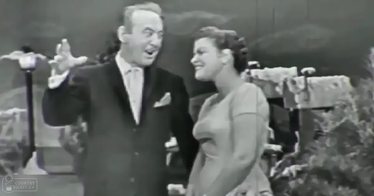 A middle-aged male singer in a dark suit with a light-colored tie holds props, while a young adult female performer in an off-white dress stands beside him on stage under bright lights.