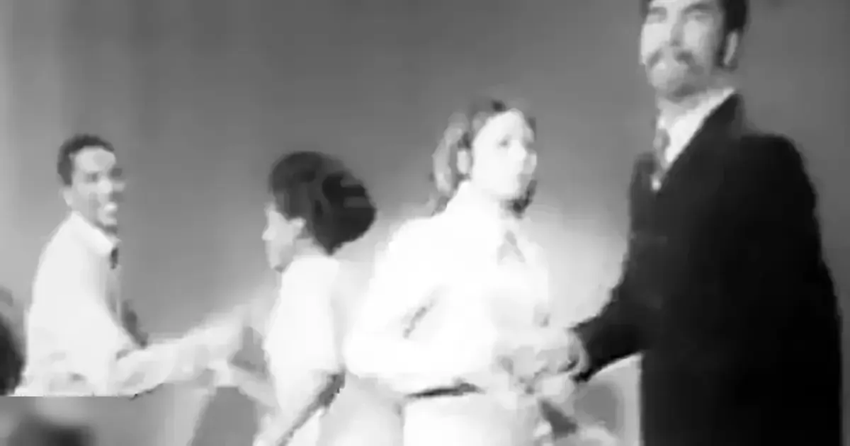 A wedding scene with an adult male singer in a light-colored suit, a young girl in a white dress with puffed sleeves, and another adult female in a white wedding dress, possibly at a church or similar venue.