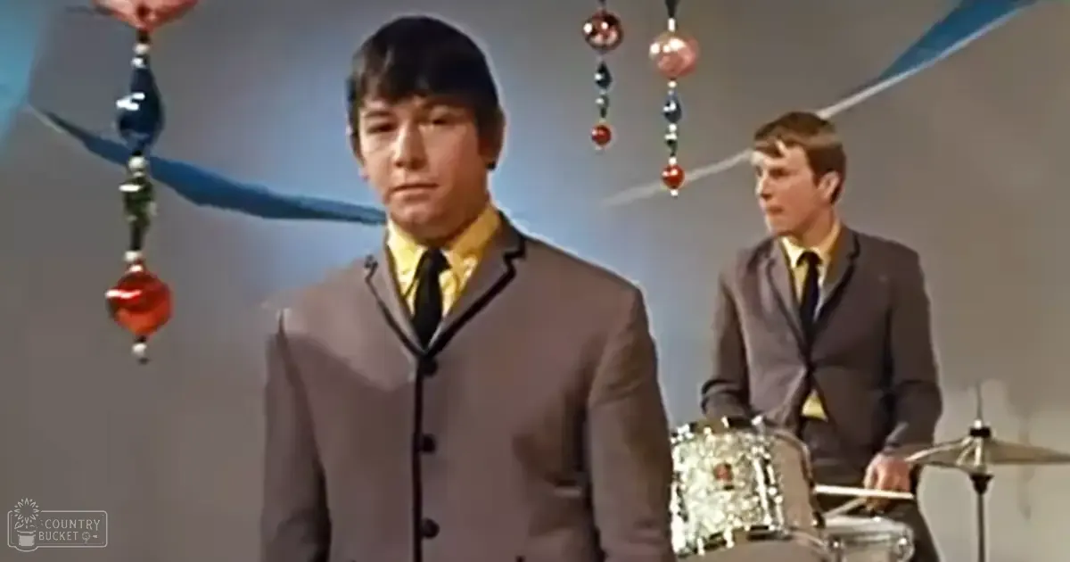 A young adult male in a gray suit jacket with black lapels and yellow shirt, standing in front of another individual playing drums; setting appears to be an indoor event or performance space.